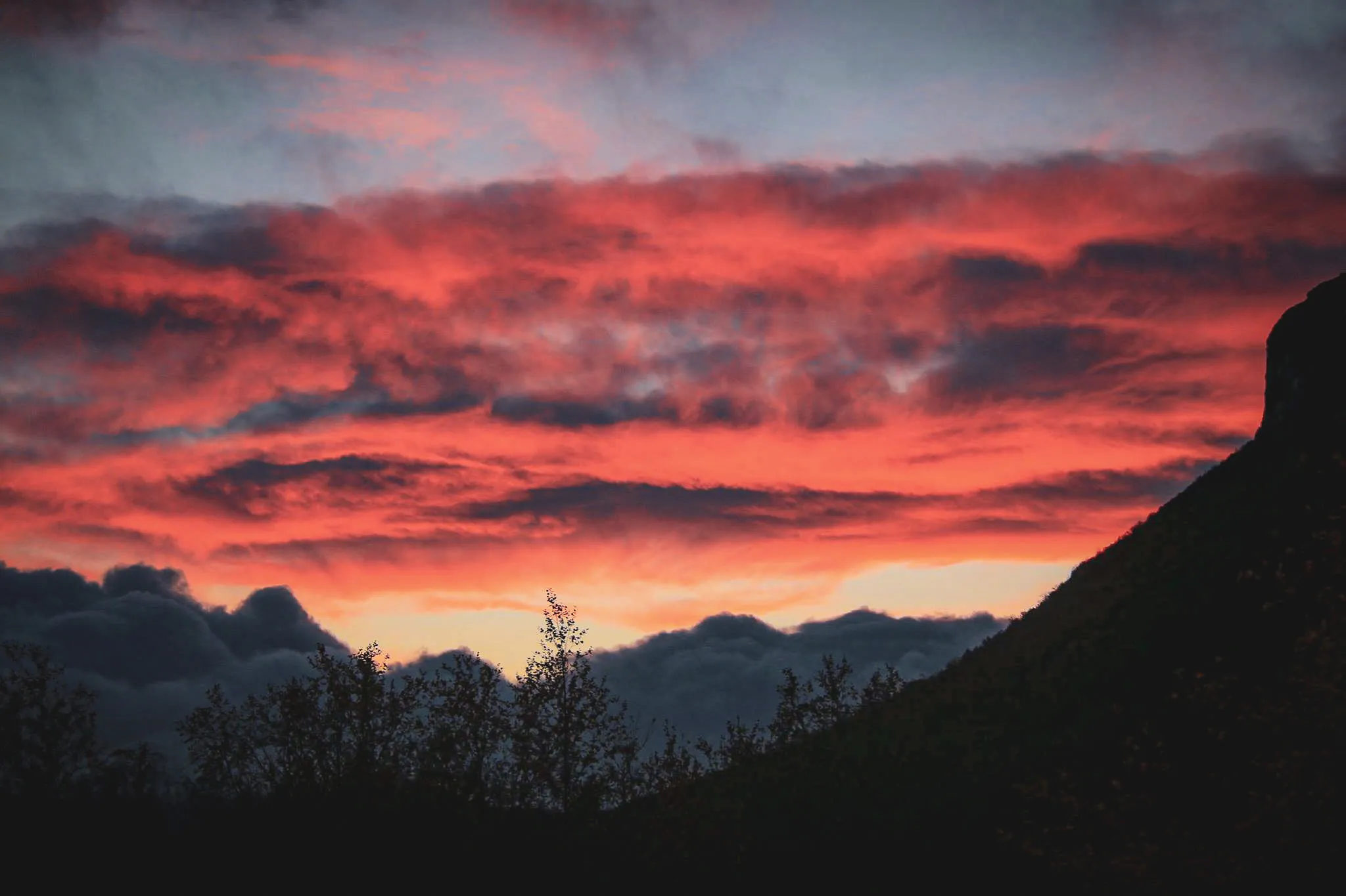 Un coucher de soleil vibrant avec des nuances de rouge, d'orange et de bleu, créant un contraste saisissant avec des nuages sombres. La silhouette d'une montagne est visible à droite.