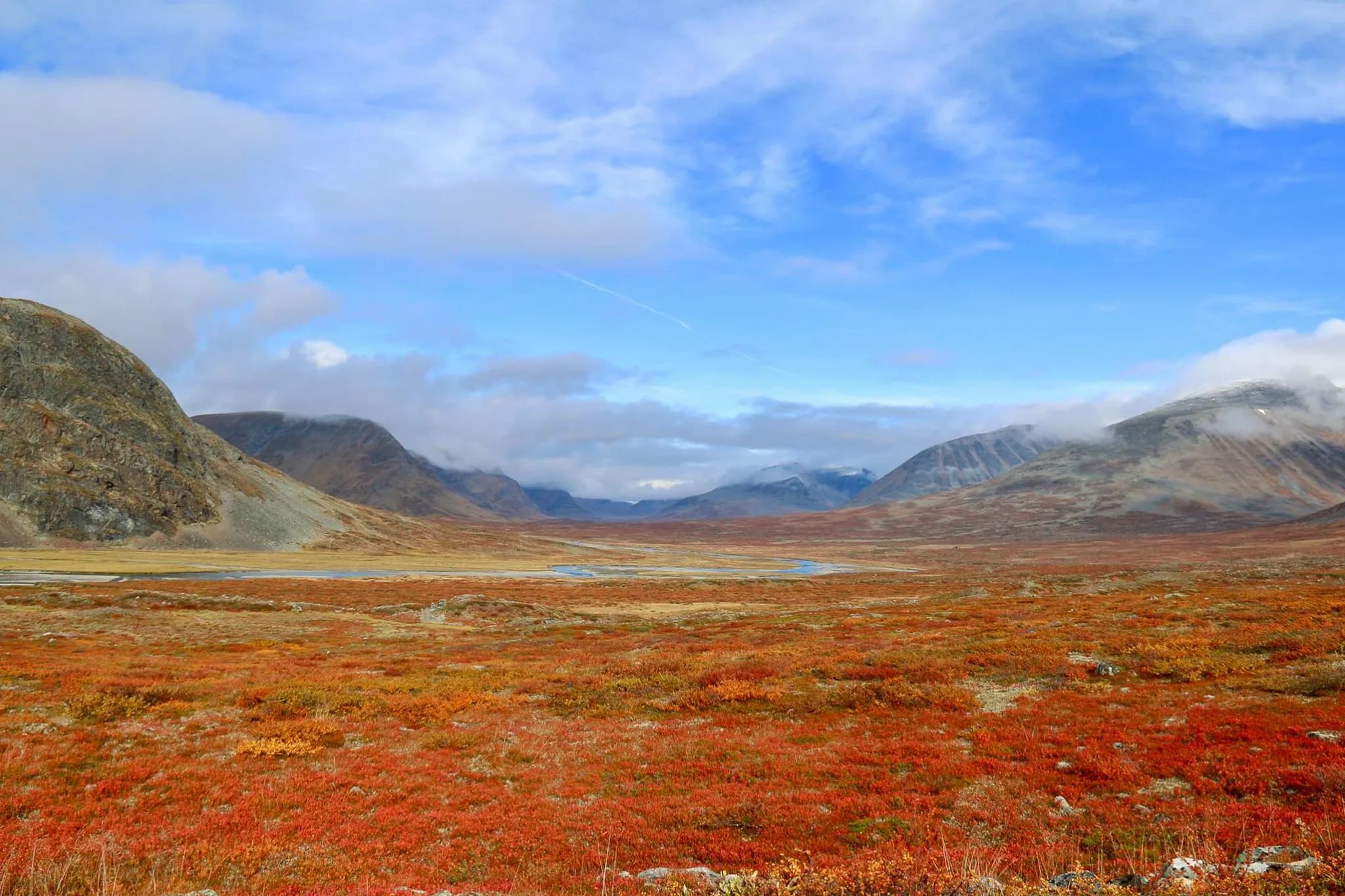 Un paysage vaste et sauvage avec des montagnes en arrière-plan, des nuages partiels dans un ciel bleu. Le sol est recouvert d'une végétation aux couleurs automnales, principalement des nuances de rouge et d'orange.
