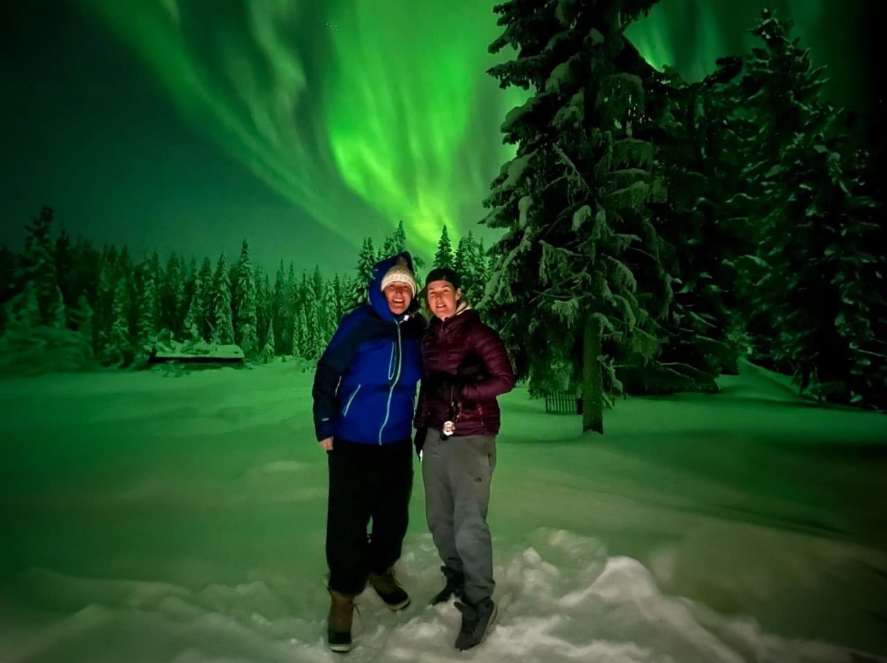 Two smiling people under the northern lights in Lapland, surrounded by snow and fir trees.