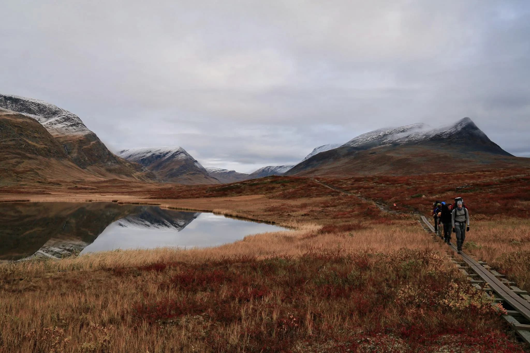 Un paysage montagneux avec des collines recouvertes de neige et des pentes douces. Un lac calme reflète les montagnes et le ciel nuageux. Dans la partie inférieure de l'image, un sentier en bois est présent.