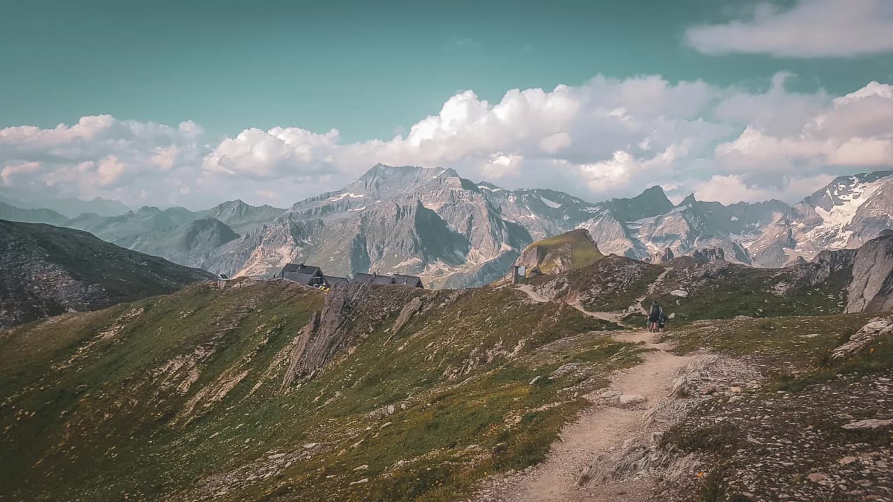 A mountain trail with huts, surrounded by majestic Alpine peaks and blue skies.