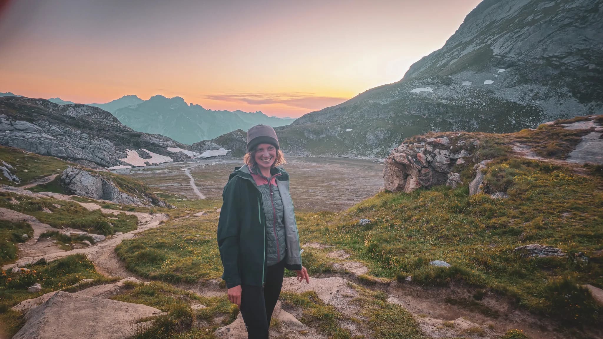 An Alpine landscape at sunset, with majestic mountains in the background. A smiling person stands on a hiking trail surrounded by greenery, wearing a jacket and hat, evoking an atmosphere of dusk.