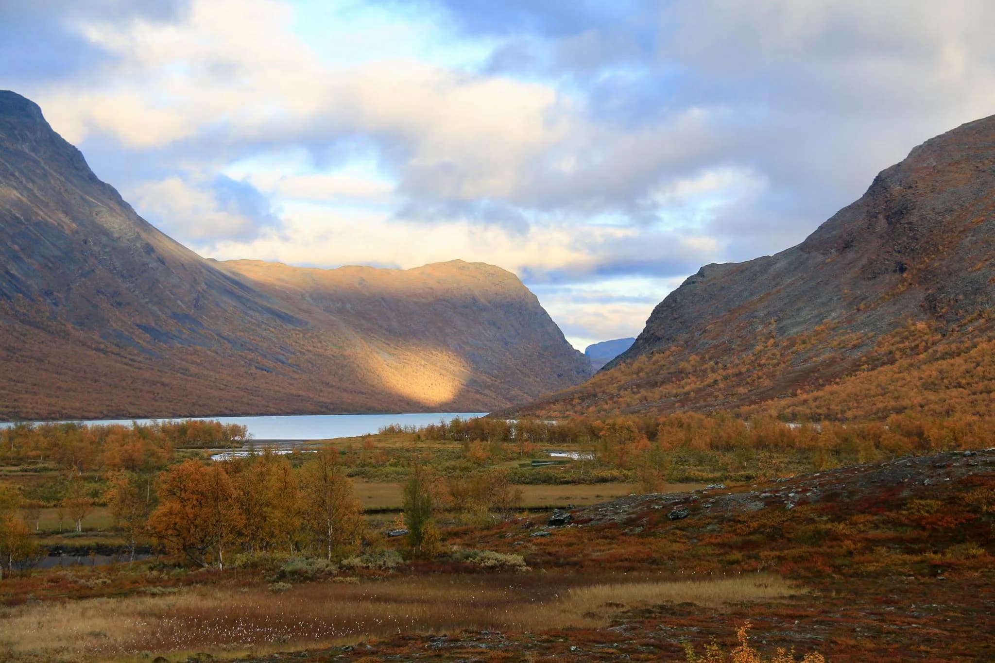 Un paysage naturel majestueux avec des montagnes encadrant une vallée. La lumière du soleil joue avec les nuages, créant des ombres douces sur les pentes rocheuses. Au fond, un lac paisible reflète le ciel.