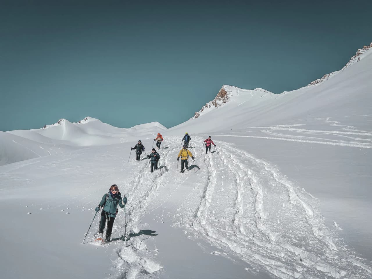 A group of snowshoe hikers on a snow-covered plateau facing majestic glaciers.