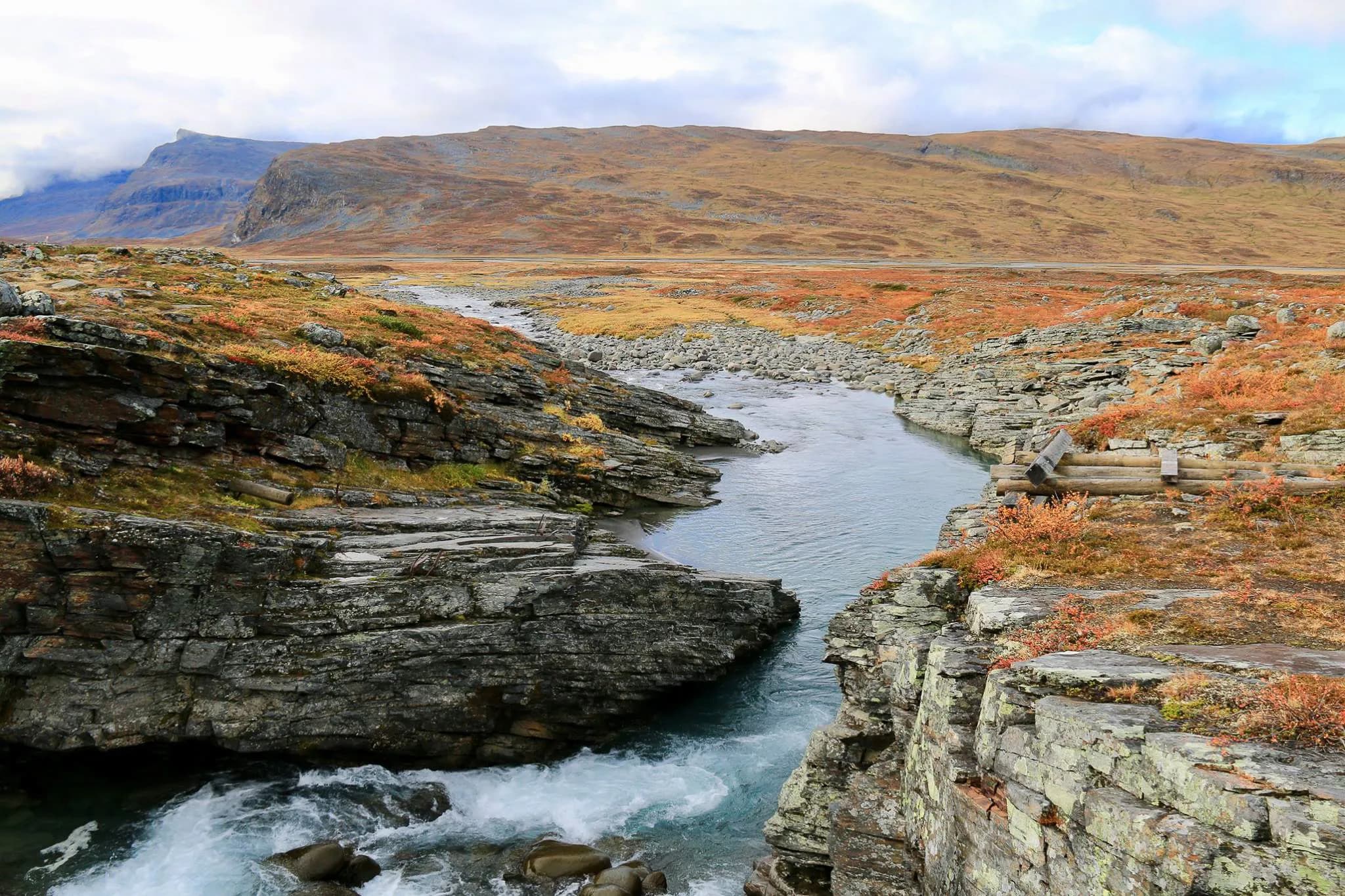 Un paysage naturel avec le cours d'eau menant à Abiskojaure serpentant entre des rochers escarpés. Les rives sont parsemées de feuillage automnal aux teintes orangées et dorées.