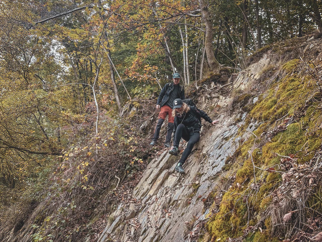 Deux randonneurs descendent un terrain escarpé en pleine forêt d'Ardenne, entourés de couleurs d'automne.