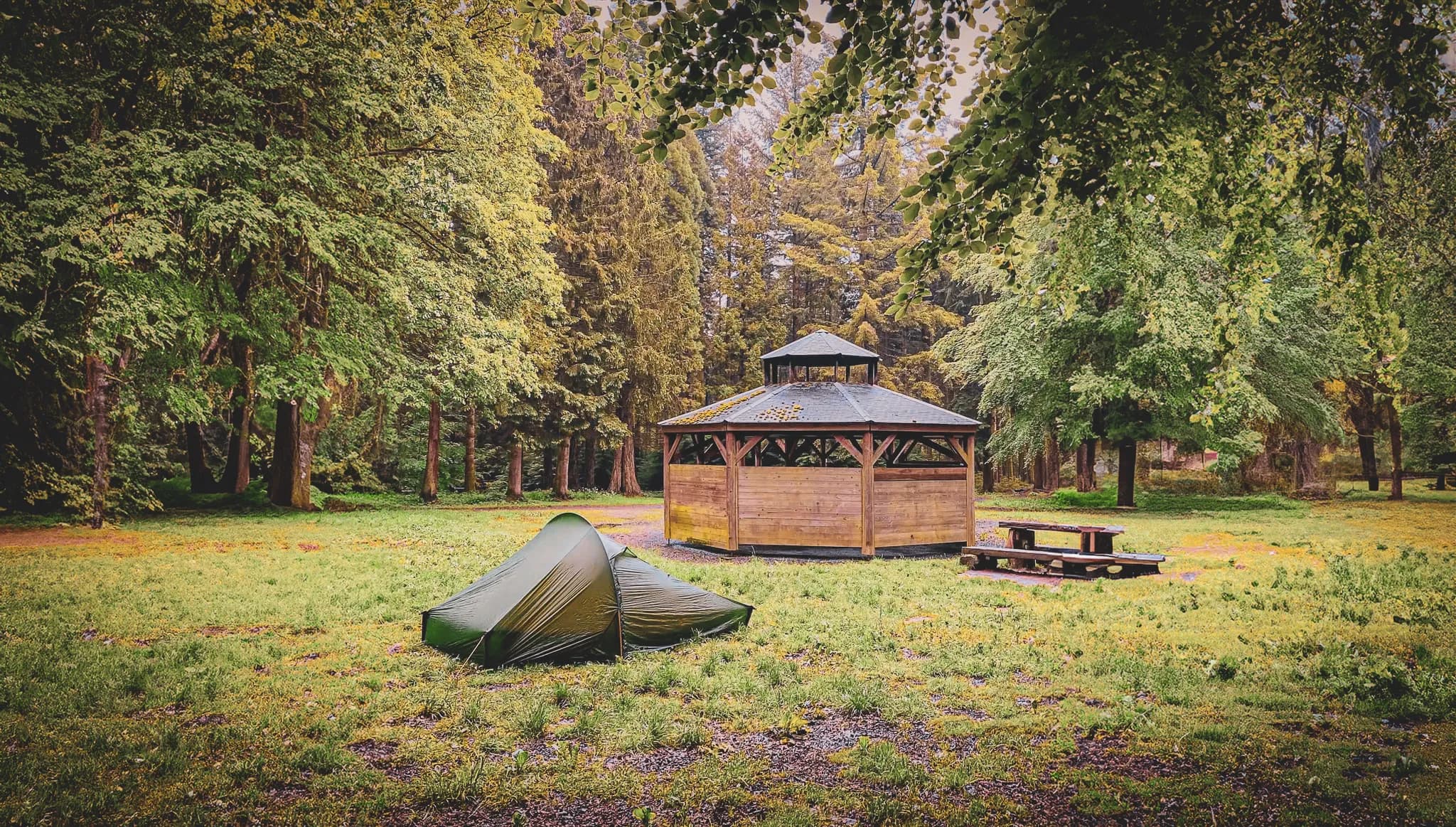 A man is sitting in front of a tent in a dense forest. He is wearing a waterproof coat and orange gloves, while examining objects in a cloth bag. Next to him, a mountain bike in the colours of
