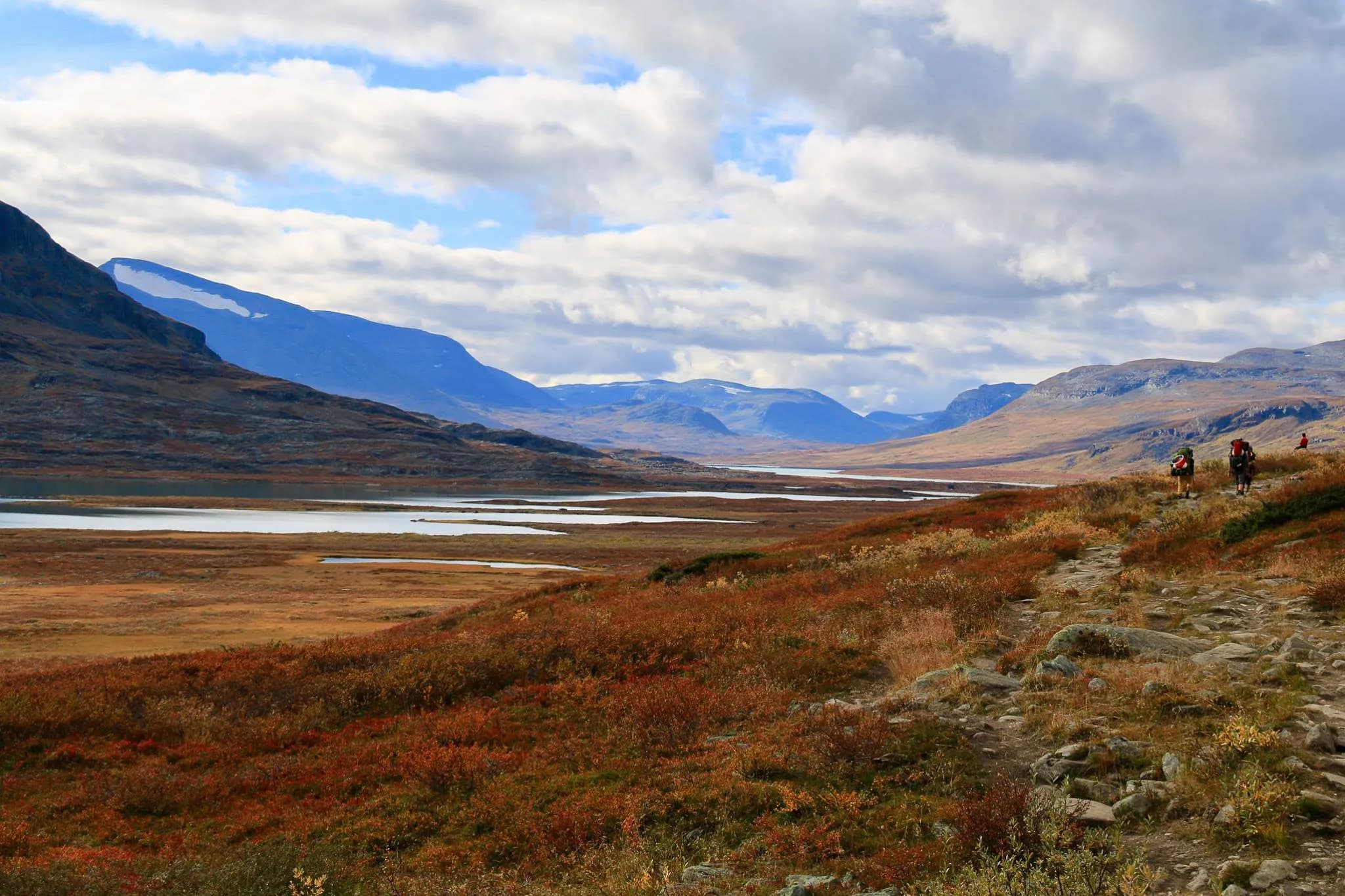 Un paysage montagneux avec des cimes majestueuses sous un ciel nuageux. Un vaste lac se reflète dans les vallées, entouré de terres aux teintes automnales.