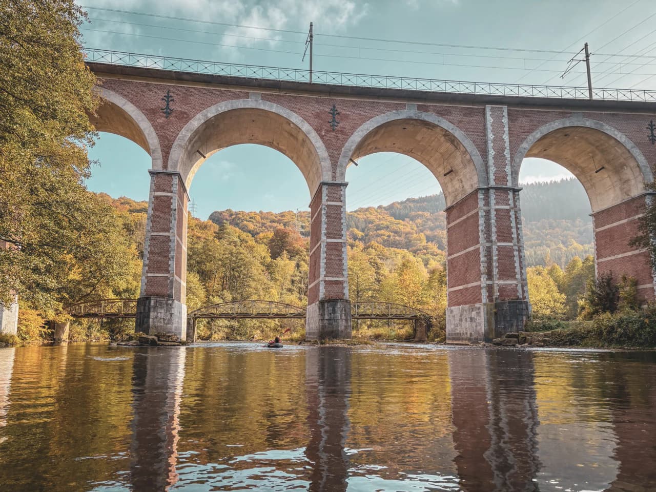 Au cœur des Ardennes, un packrafteur navigue sous un majestueux viaduc, entouré de couleurs automnales.