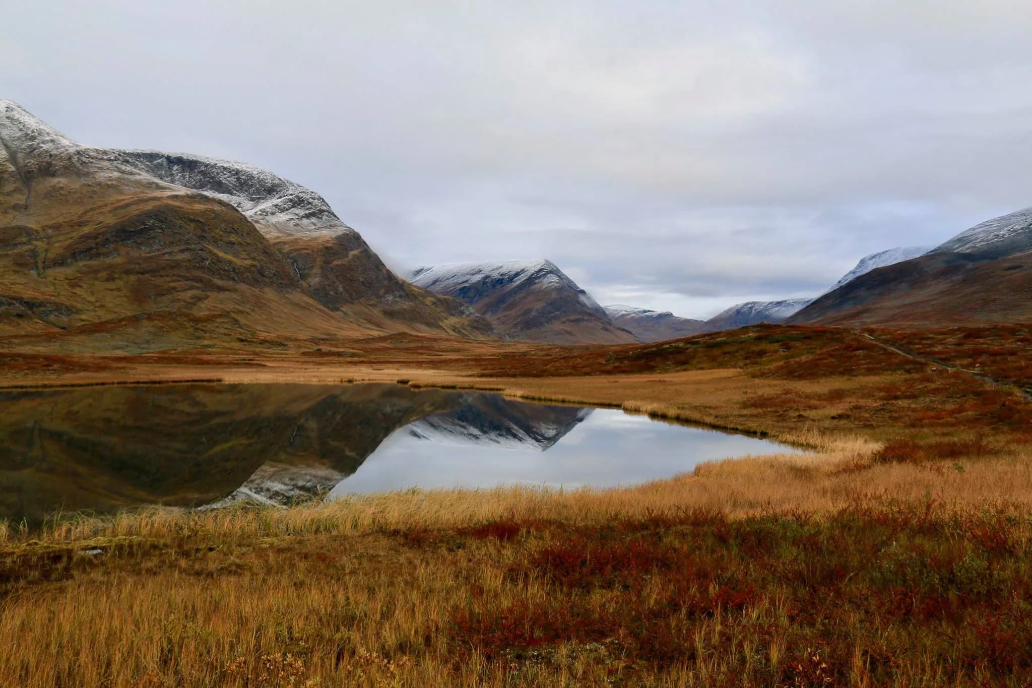 Un paysage montagneux au coucher du soleil, avec des sommets enneigés et une vaste plaine recouverte d'herbes dorées. Un lac calme reflète les montagnes environnantes, créant une harmonie entre les éléments naturels.