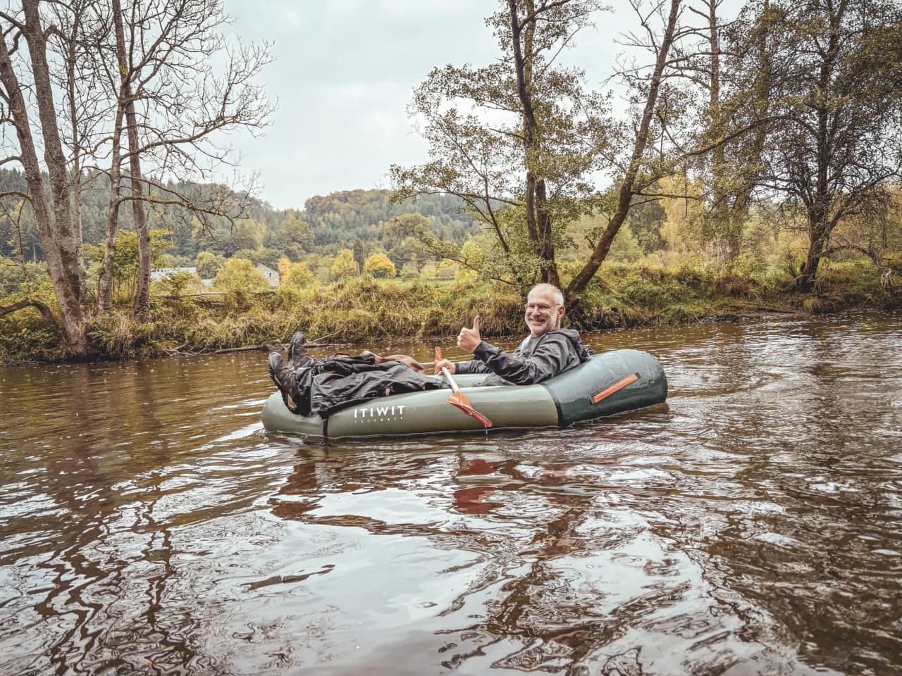 Un aventurier souriant dans un packraft, entouré de paysages verdoyants en Ardenne.