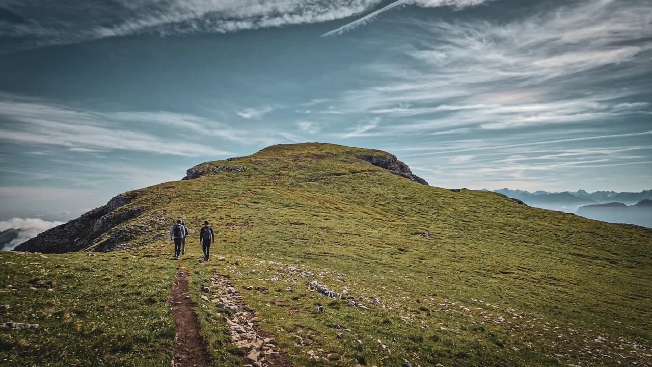 Two hikers are walking along a leafy path leading to Mont Aiguille, under a clear sky.