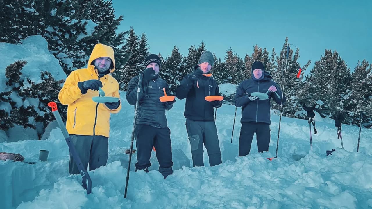 Four adventurers on snowshoes enjoy a meal in the snow, facing the mountains.