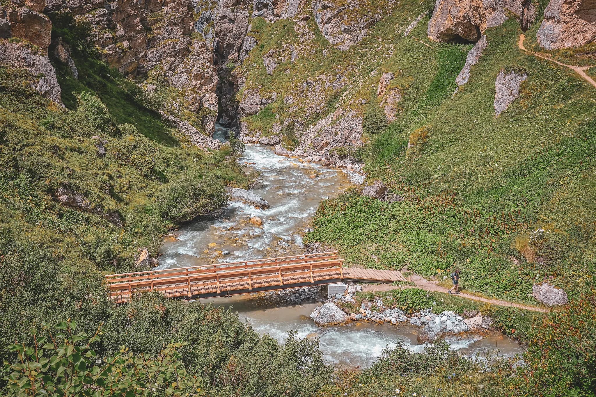 A spectacular mountain panorama reveals majestic peaks with jagged edges. In the foreground, a winding path crosses green slopes dotted with rocks. To the left, a number of
