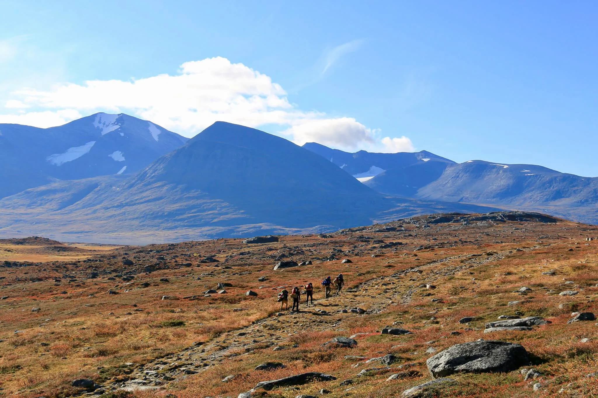 Un paysage montagneux dans une région sauvage avec des collines et des montagnes en arrière-plan, partiellement recouvertes de neige. Le ciel est d'un bleu clair, parsemé de nuages blancs.