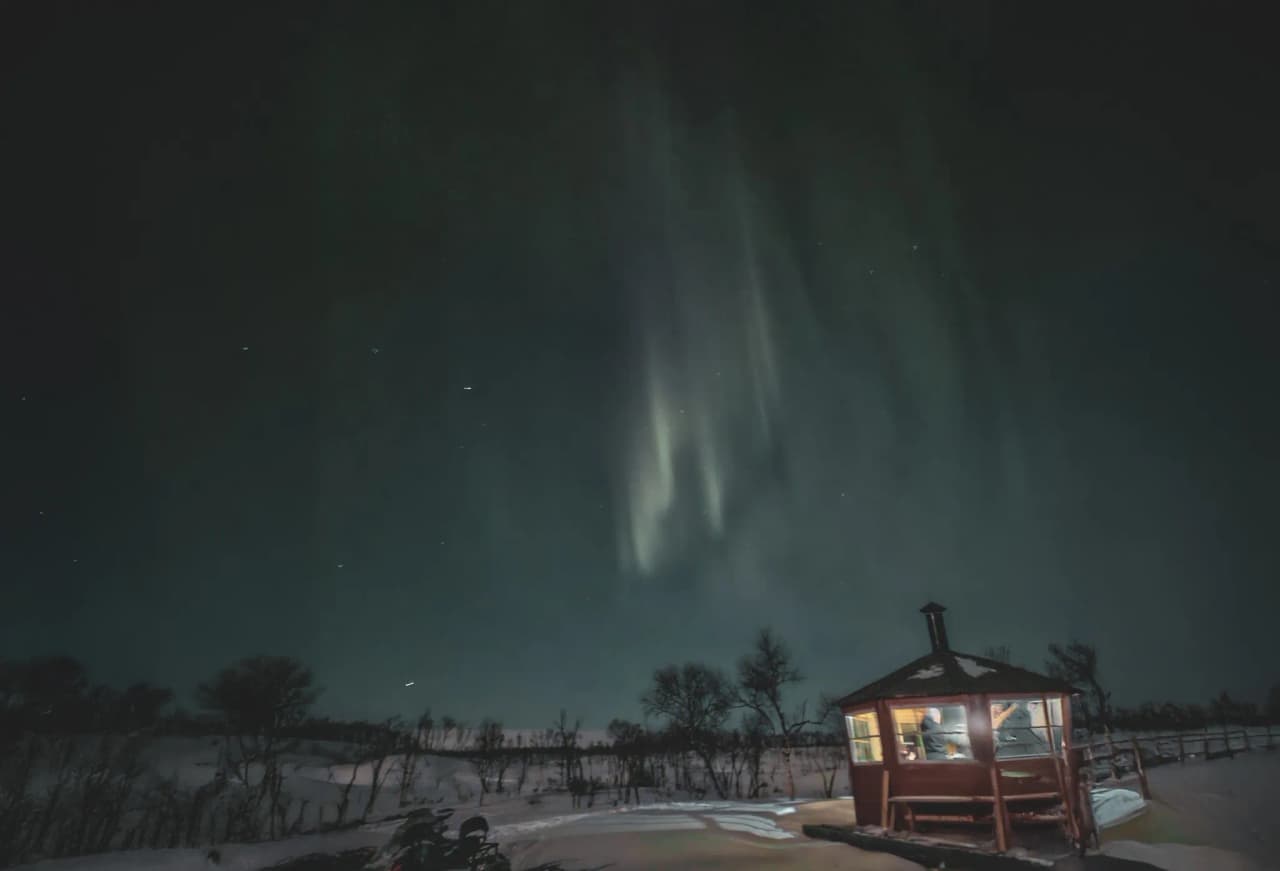 An illuminated cabin under the northern lights, surrounded by snow, an invitation to adventure in Lapland.