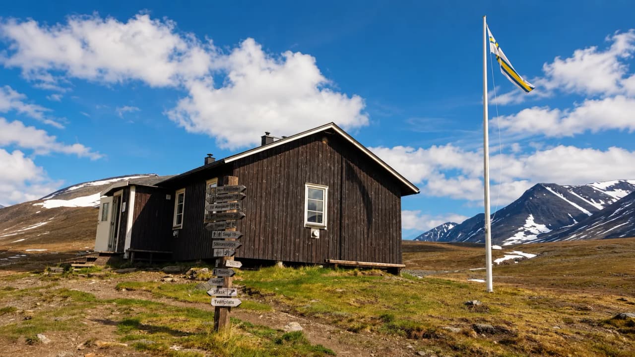 Cabane en bois typique sous un ciel bleu, entourée de montagnes enneigées en Laponie suédoise.